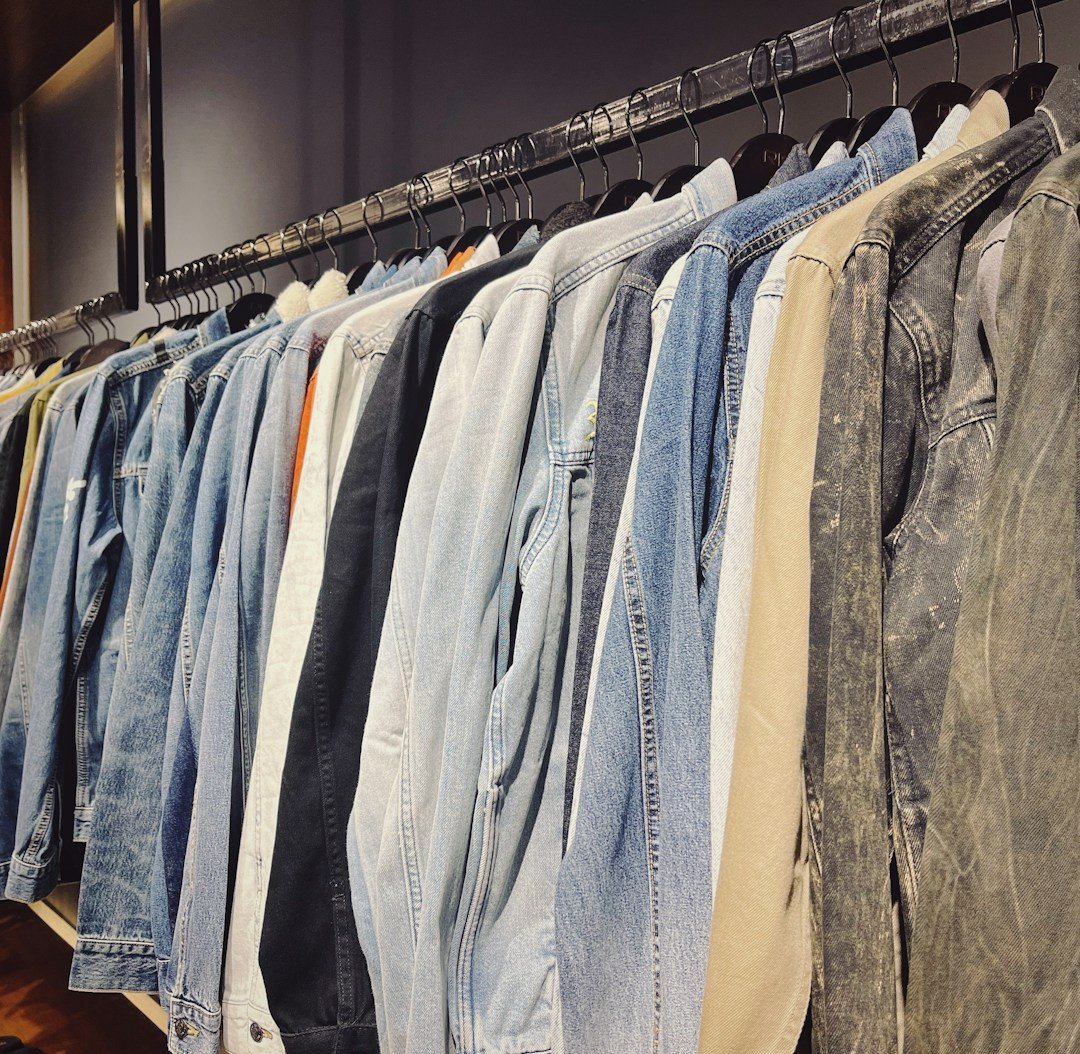A neatly organized row of jackets hanging on a clothing rack, featuring a variety of denim, tan, and neutral colors. The wooden wall and shelves add a warm touch to the setting, with a few pairs of sandals placed below.